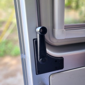 May include: A black window winder with a silver handle is mounted on a silver vehicle door. The winder features a small alien head design. The window frame is white.
