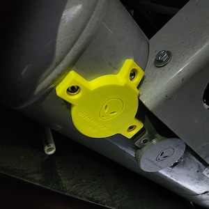 May include: A yellow, alien-themed cover with the text "alienenterprise.net" and a gray, alien-themed cap are attached to a gray metal structure. The cover is secured with screws.