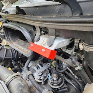 May include: A close-up view of a car engine compartment. A bright red rectangular metal bracket is bolted to a silver metal frame. The engine components include black hoses, pipes, and other mechanical parts.