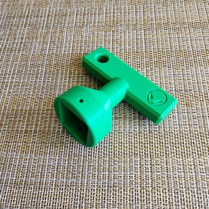 May include: Bright green, 3D-printed tool with an alien head logo. The tool has a rectangular handle with a hole and a square-shaped head. The tool is designed for a specific purpose, possibly for crafts or DIY projects.