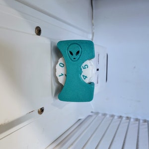 May include: A teal-colored, 3D-printed alien-themed refrigerator dial cover. The cover is shaped to fit over the dial, with an alien face design at the top. The dial has numbers 0-5.