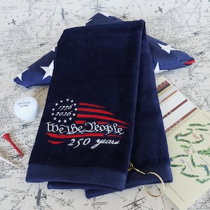 May include: Navy blue golf towel with embroidered American flag design and the text "We the People 250 years". Includes a golf ball, tee, and scorecard. The towel has a loop for hanging.