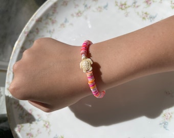 Puede incluir: Un brazalete de cuentas de colores con un dije de tortuga blanca. El brazalete está hecho de cuentas rosas, naranjas y blancas.