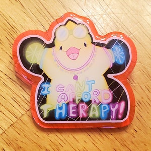 May include: A colorful, star-shaped pin with a cartoon character wearing goggles and a necklace. The pin has the text "I CAN'T AFFORD THERAPY!" in vibrant, multi-colored letters, outlined in black and red.