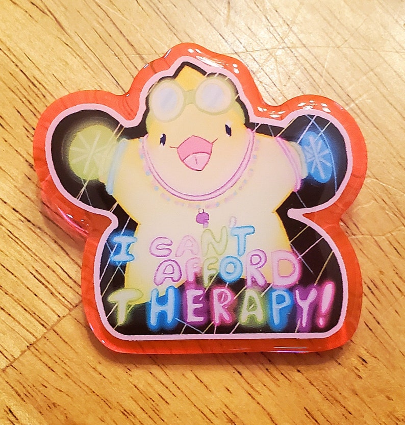 May include: A colorful, star-shaped pin with a cartoon character wearing goggles and a necklace. The pin has the text "I CAN'T AFFORD THERAPY!" in vibrant, multi-colored letters, outlined in black and red.