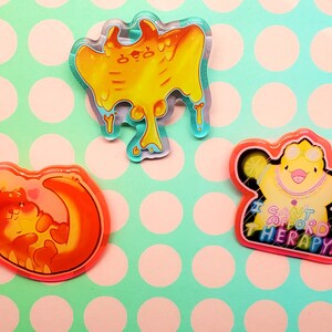 May include: Three colorful acrylic pins. One is a yellow blob with dripping accents. Another is an orange otter curled around a crescent moon. The third is a yellow duck with the text "I CAN'T AFFORD THERAPY!"