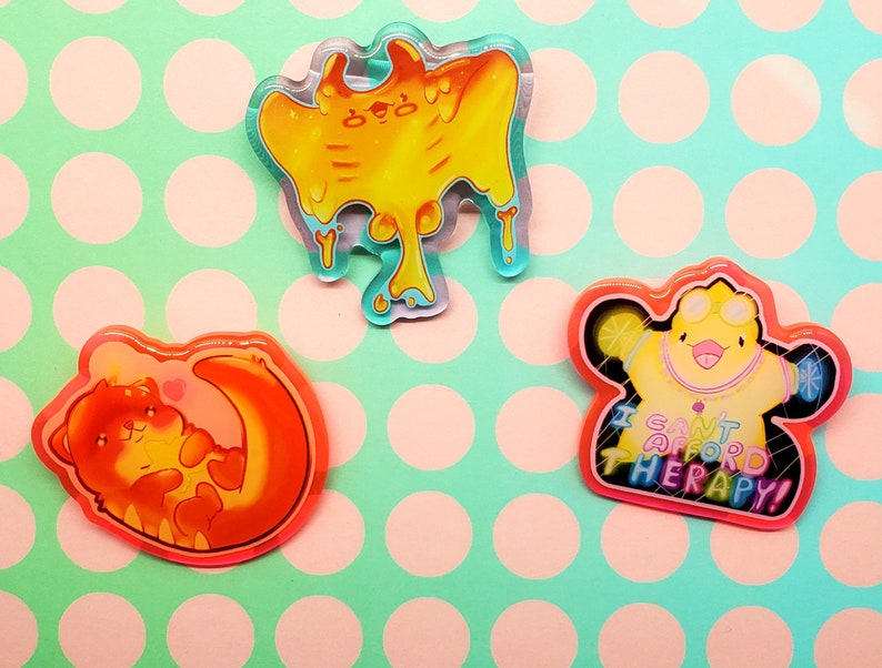 May include: Three colorful acrylic pins. One is a yellow blob with dripping accents. Another is an orange otter curled around a crescent moon. The third is a yellow duck with the text "I CAN'T AFFORD THERAPY!"