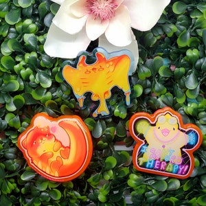 May include: Three colorful acrylic pins with unique designs. One pin features a yellow blob with blue accents, another shows a sleeping bear with hearts, and the third displays a chick with text that reads "I CAN'T AFFORD THERAPY!"