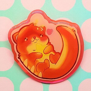 May include: A sticker featuring a cartoon otter in shades of orange and yellow, holding a star-shaped object. The otter is surrounded by a pink outline with a heart, set against a pink and blue polka dot background.