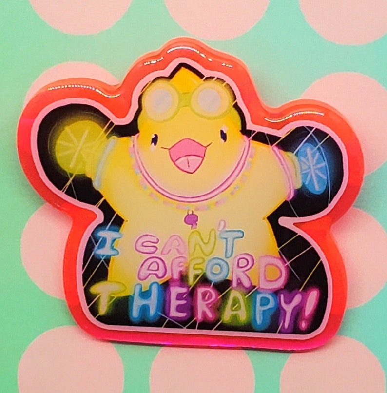 May include: A colorful pin featuring a cartoon character with goggles and a necklace. The character is yellow with a pink tongue and is inside a red and pink outline. The text "I CAN'T AFFORD THERAPY!" is written across the bottom.