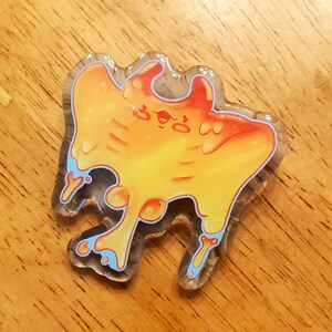 May include: A clear acrylic pin with a cartoon design. The pin features a yellow, orange, and red creature with blue accents. The pin has a unique, organic shape and is set against a wooden background.