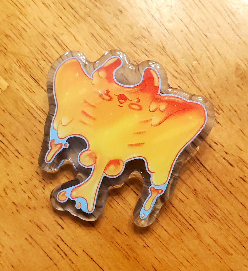 May include: A clear acrylic pin with a cartoon design. The pin features a yellow, orange, and red creature with blue accents. The pin has a unique, organic shape and is set against a wooden background.