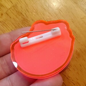 May include: A neon orange pin with a white safety clasp and a silver pin. The pin is in the shape of a flower. The pin is made of a clear, glossy material.