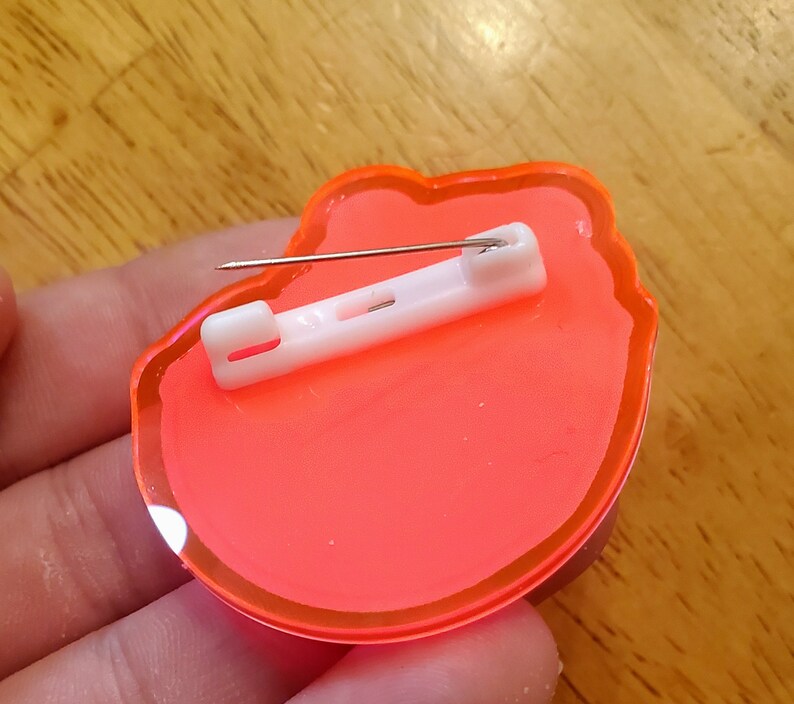 May include: A neon orange pin with a white safety clasp and a silver pin. The pin is in the shape of a flower. The pin is made of a clear, glossy material.