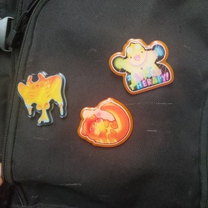 May include: Three colorful enamel pins are attached to a black backpack. One pin features a yellow creature with the text "I CAN'T AFFORD THERAPY!". The other two pins have orange and yellow designs.
