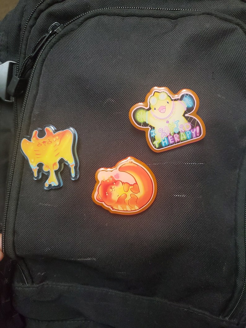 May include: Three colorful enamel pins are attached to a black backpack. One pin features a yellow creature with the text "I CAN'T AFFORD THERAPY!". The other two pins have orange and yellow designs.
