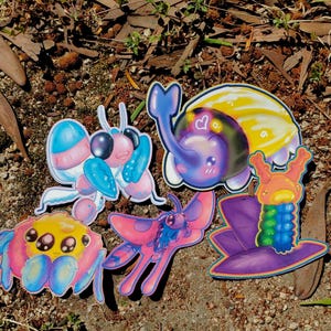 May include: A collection of colorful, cartoon-style stickers featuring various insect-like creatures. The stickers showcase a range of vibrant colors, including pink, blue, yellow, and purple. The designs include a spider, a moth, and a beetle.
