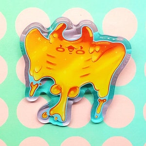 May include: A colorful, cartoonish acrylic pin featuring a stylized, orange and yellow creature with a dripping, melted appearance. The pin has a teal outline and a clear, glossy finish. The design is whimsical and playful.