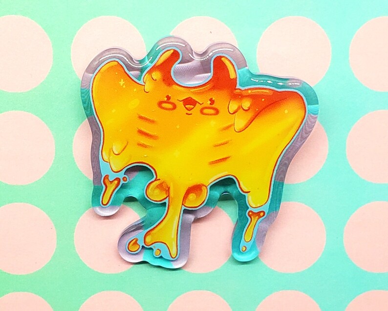 May include: A colorful, cartoonish acrylic pin featuring a stylized, orange and yellow creature with a dripping, melted appearance. The pin has a teal outline and a clear, glossy finish. The design is whimsical and playful.