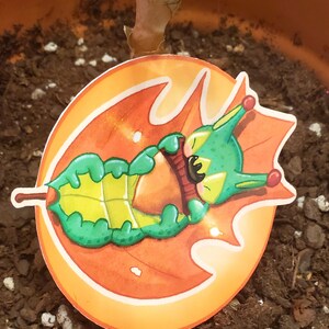 May include: A sticker featuring a cartoon creature resting on a stylized orange leaf. The creature is green and yellow with red accents. The leaf has a brown stem and is set against a light orange background.