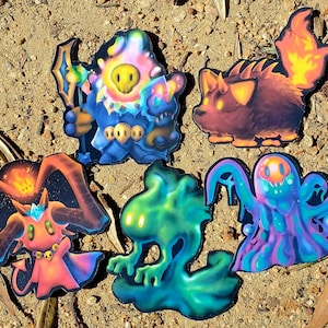 May include: A collection of five colorful fantasy creature stickers. The stickers feature various designs, including a blue wizard, a fiery fox-like creature, a pink and orange demon, a green blob, and a multi-colored ghost. The stickers are on a sandy surface.