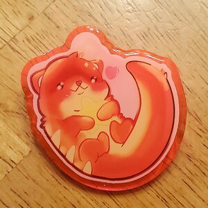 May include: A colorful, cartoon-style sticker featuring an otter holding a heart-shaped object. The otter is primarily orange with white accents, set against a pink background with a heart. The sticker has a glossy finish and a clear border.