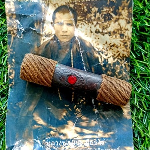 Może przedstawiać: Brązowo-czarny amulet z czerwonym klejnotem w środku, owinięty brązowym sznurkiem. Amulet leży na czarno-białym zdjęciu młodego mężczyzny.