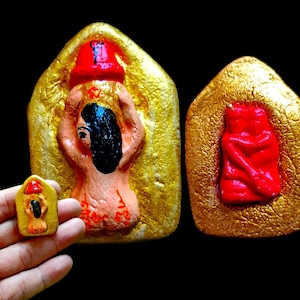 Può includere: Tre amuleti dorati con figure dipinte in rosso e nero. Gli amuleti hanno la forma di una casa, un rettangolo e una casa più piccola. Le figure sono stilizzate e astratte.