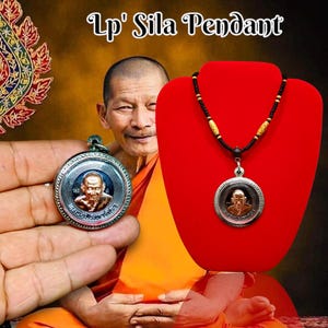May include: A silver pendant necklace with a round silver charm featuring a portrait of a Buddhist monk. The charm is on a black cord with brown wooden beads. The pendant is displayed on a red velvet stand.