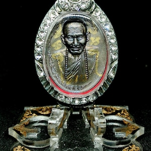 Pode incluir: Um pingente de amuleto de prata e ouro com o retrato de um monge com um colar de contas. O pingente é cercado por strass transparentes e é exibido em um suporte de acrílico transparente com detalhes dourados.