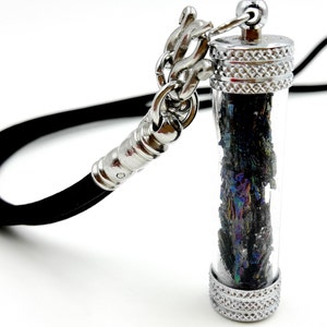 Può includere: Una collana con un ciondolo in argento e un tubo di vetro trasparente riempito di cristalli neri iridescenti. La collana ha un cordoncino nero e una chiusura in argento.