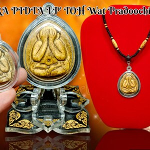 May include: A gold amulet pendant with a Buddha image, on a black and brown cord necklace. The amulet is in a clear plastic case, and the necklace is on a red velvet background. The text "PRA PIDTA LP TOJH Wat Pradoochimplee" is visible at the top of the image.
