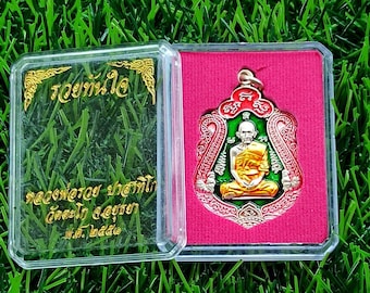 Puede incluir: Un colgante de amuleto dorado y verde con una figura de Buda sentado en un marco adornado rojo y blanco. El colgante está en una caja de plástico transparente con interior rosa. La caja tiene texto tailandés.