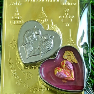 May include: A gold-colored metal frame with two heart-shaped charms. The left charm is silver and features two birds with wings outstretched. The right charm is red and features two birds with wings outstretched.