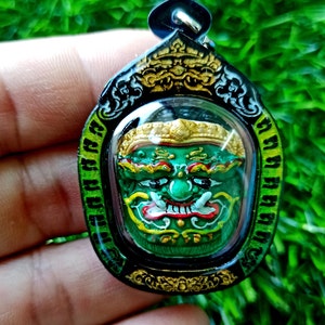 May include: A green and gold amulet featuring a detailed face with a fierce expression. The amulet is framed in black and has a gold trim. The amulet is on a hand.