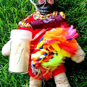 May include: A small, red fabric doll with a black skull head and pink eyes. The doll is wearing a colorful rope necklace and has a small glass jar with white powder next to it.