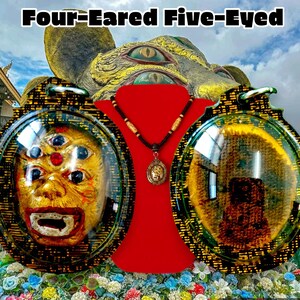 May include: Two oval-shaped amulets with gold and black patterned frames. The amulet on the left has a glass dome revealing a golden face with five eyes and a wide open mouth. The amulet on the right has a glass dome revealing a dark brown image. The amulets are displayed on a red velvet surface with a wooden beaded necklace and a small pendant.