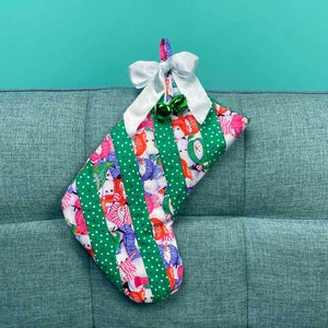 以下が含まれることがあります： 雪だるまやその他の冬のモチーフが描かれた緑と白のクリスマスストッキング。ストッキングには白いリボンと緑色の鈴が2つ付いています。