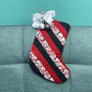 以下が含まれることがあります： ペンギン柄の赤と黒のストライプのクリスマスストッキング。ストッキングには白いリボンと2つの銀色のベルが付いています。ストッキングは青いソファに掛けられています。