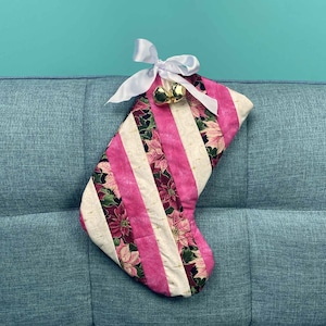 以下が含まれることがあります： ピンクと白のストライプのクリスマスストッキングに、花柄と金のベルが上部にぶら下がっています