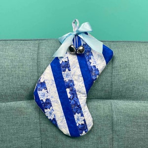 以下が含まれることがあります： 青と白のクリスマスストッキング。青と白の花柄と、銀色のベルが付いた青いリボンが付いています。