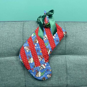 以下が含まれることがあります： 手作りのクリスマスストッキング。赤と青の生地に白い水玉模様と犬のイラストがパッチワークされています。緑色のサテンリボンと小さなベルでトリミングされています。