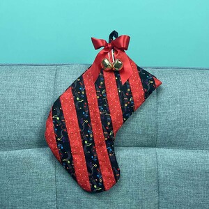 以下が含まれることがあります： 音楽のモチーフと赤いリボンと金のベルが付いた赤と黒のクリスマスストッキング。ソファに掛けられています。