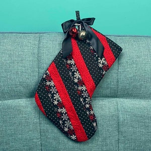 以下が含まれることがあります： 青いソファに、白の雪の結晶模様の入った赤と黒のクリスマスストッキングが掛けられています。ストッキングには黒いリボンと銀色のベルの飾りがあります。