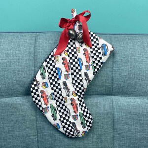 以下が含まれることがあります： 赤と白のチェック柄のクリスマスストッキングで、カラフルな車のデザインが施されています。ストッキングの上部には赤いリボンと鈴が付いています。