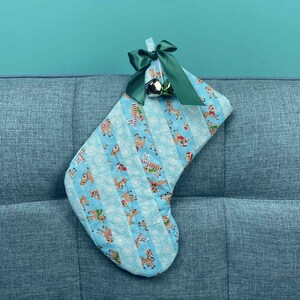 以下が含まれることがあります： 緑のリボンと銀のベルが付いた青と白のクリスマスストッキング。ストッキングはトナカイの模様で飾られています。