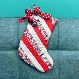 以下が含まれることがあります： お祝いのペンギンの模様が施された手作りのクリスマスストッキング。ストッキングは赤と白のストライプ、赤い水玉模様のリボン、銀色のベルが特徴です。灰色の布張りの表面に置かれています。