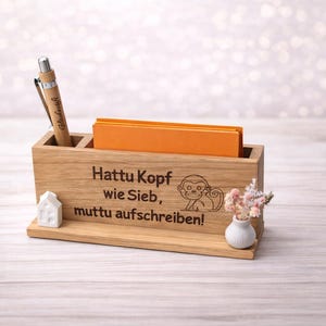 Könnte beinhalten: Ein rechteckiger Schreibtisch-Organizer aus Holz mit Stiftehalter, einem Schlitz für orangefarbene Notizzettel und einer kleinen weißen Hausfigur. Die Vorderseite ist mit einer Affen-Illustration und dem Text Hattu Kopf wie Sieb, muttu aufschreiben! graviert.