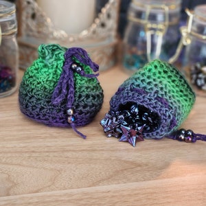 May include: Two crocheted drawstring bags in shades of green and purple with a purple and black beaded drawstring. One bag is filled with iridescent purple and black star-shaped dice.