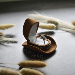 May include: A turquoise ring with silver accents sits inside a brown suede ring box shaped like a cowboy hat. The box is open and the ring is visible. The background is a light grey surface with dried grasses.